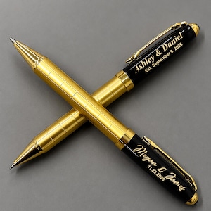 May include: Two ballpoint pens with gold-toned grid-patterned barrels and black caps. One pen is engraved with Ashley & Daniel Est. September 9, 2026, and the other with Megan & James 11.23.2026. Both pens feature gold-colored clips and tips.