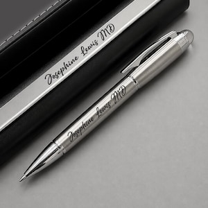 May include: A silver-toned ballpoint pen and a matching silver-toned pen case, both engraved with the name Josephine Lewis MD in a black script font. The pen features a polished metal clip and a smooth barrel, while the case has a black finish.