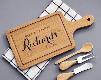 May include: A rectangular bamboo serving board with a handle, engraved with the text Ryan & Jennifer Richards est. 2026. The set includes a small wooden spoon, a two-pronged metal fork with a wooden handle, and a metal spreader with a wooden handle.