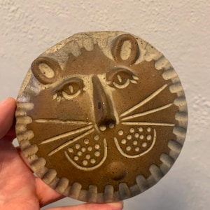 May include: A round, ceramic lion face with a textured, brown finish. The lion has detailed eyes, a nose, and whiskers. The rim of the ceramic piece has a decorative, ridged edge. The lion face is a decorative item.