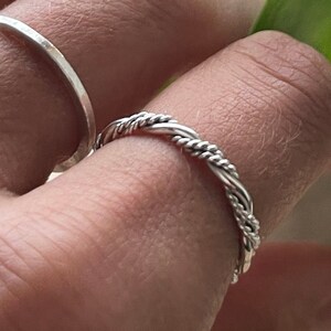 May include: Two silver rings on a finger. The ring on the index finger is a thin band. The ring on the middle finger is a twisted band.
