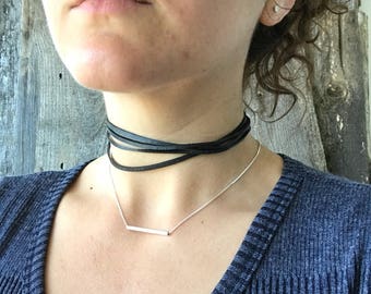 May include: A woman wearing a black leather choker necklace and a silver bar necklace. She is wearing a blue sweater.