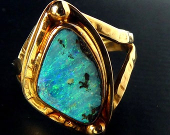 May include: A gold ring featuring a large, iridescent opal stone. The opal displays vibrant blues and greens with dark inclusions. The ring has a thick, textured gold band and a triangular setting that frames the stone.
