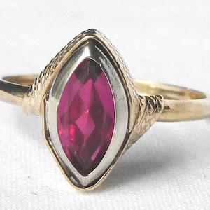 May include: A gold ring with a marquise-shaped ruby gemstone set in a white metal bezel. The ring band is decorated with a twisted design.