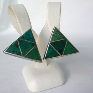 May include: A pair of silver triangle earrings with green gemstone inlay. The earrings are on a white display stand.