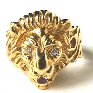 May include: A gold-colored lion ring with a detailed mane and face. The lion has two small diamonds for eyes and a small purple stone at the mouth. The ring is a statement piece.