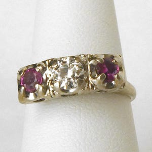 May include: A gold ring with three stones. The center stone is a clear diamond, and the two stones on either side are pink rubies.