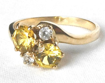 Könnte beinhalten: Ein goldfarbener Ring mit einem Cluster aus gelben und klaren Edelsteinen. Der Ring hat ein geschwungenes Band und eine Ansammlung von drei gelben Steinen und zwei klaren Steinen. Die Edelsteine sind in einem goldfarbenen Metall gefasst.