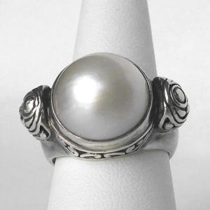 May include: A silver ring with a large white pearl set in the center. The ring has a detailed design with two smaller pearl-like accents on either side of the main pearl.