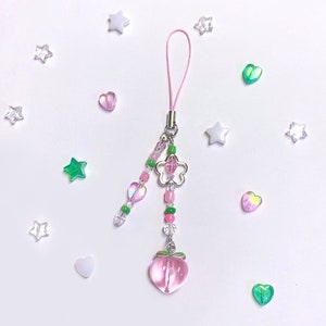 Pode incluir: Um amuleto de pêssego rosa com um amuleto de flor prateado e contas verdes e rosa em um cordão rosa. O amuleto está preso a uma alça de telefone rosa.