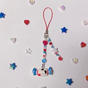 Puede incluir: Un charm de teléfono con cuentas rojas, blancas y azules con un charm con forma de caramelo que presenta una cara de dibujos animados. El charm está unido a una cuerda roja.
