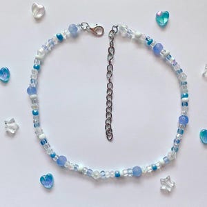 May include: A delicate choker necklace featuring a mix of blue, white, and clear beads. The necklace has a silver-toned chain and clasp, with a beaded extension. Decorative heart and star-shaped beads surround the necklace.