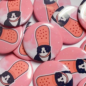 Può includere: Una collezione di spille a bottone rosa con gatti neri e bianchi che indossano cappelli rosa a forma di cerotto. I gatti hanno pelliccia bianca intorno agli occhi e una macchia bianca sul petto.