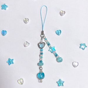 May include: A light blue phone charm with a heart-shaped pendant, star beads, and a round bead. The charm is made with silver-toned metal and blue glass beads.