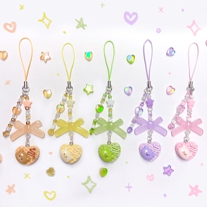 Könnte beinhalten: Fünf bunte Handy-Anhänger mit herzförmigen Anhängern. Jeder Anhänger hat ein anderes Farbschema, darunter Pfirsich, Gelb, Grün, Lila und Rosa. Die Anhänger haben Perlen, Schleifen und sternförmige Details.