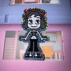 May include: A pixelated enamel pin featuring a woman with dark hair, wearing a black top and pants. The pin has a glittery, iridescent border. The design is reminiscent of 8-bit video game graphics.