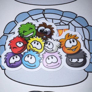 May include: A sticker featuring a group of ten colorful, round, furry creatures with large eyes and expressive faces, including red, green, brown, yellow, white, orange, purple, blue, pink, and black characters, set against a light blue igloo background.