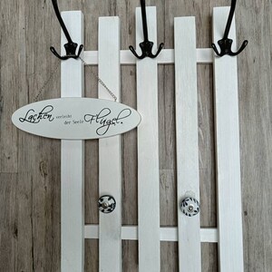 Op de afbeelding: Een witte houten kapstok met drie zwarte haken aan elke kant. De kapstok is gevormd als een schutting. Een wit ovaal bord hangt aan de bovenkant met de tekst "Lachen verleiht der Seele Flügel."