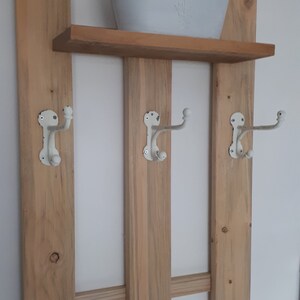 Könnte beinhalten: Ein Wandregal aus Holz mit drei Haken und einem kleinen weißen Topf oben. Das Regal hat einen rustikalen Look mit einer natürlichen Holzoptik. Die Haken sind aus weißem Metall mit einer patinierten Oberfläche. Der Topf ist oval geformt mit einem erhabenen Design.
