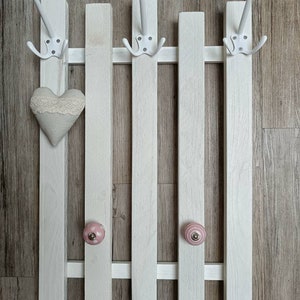 Op de afbeelding: Een witte houten kapstok met vijf haken en twee roze keramische knoppen. De kapstok is gemonteerd op een houten muur.