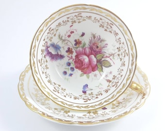 May include: A white teacup and saucer set with a floral design. The teacup features a large pink rose surrounded by other flowers in shades of purple, blue, and yellow. The rims are scalloped and trimmed with gold.