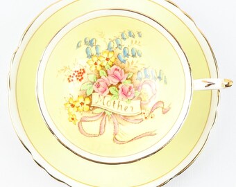 May include: A yellow teacup and saucer set with a floral design and the word "Mother" on a banner. The floral design features pink roses, bluebells, and yellow flowers.