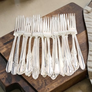 May include: A collection of antique silver forks arranged on a dark wooden cutting board. The forks have ornate handle designs and are displayed in a fan-like pattern. A neutral-colored cloth is in the background.