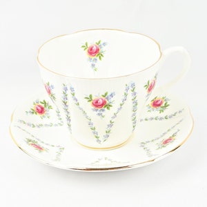 Peut inclure: Une tasse à thé et une soucoupe blanches avec un motif floral délicat représentant des roses roses et des myosotis bleus. La tasse et la soucoupe ont une bordure dorée.