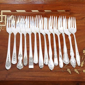 May include: A collection of silver forks with ornate handle designs, arranged on a dark wooden surface with decorative inlays. The forks vary in pattern, showcasing floral and geometric motifs.
