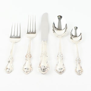 May include: A collection of five silver-coloured cutlery items, comprising two forks, a knife, and two spoons, displayed on a white surface. The silverware has elaborate floral patterns on the handles, indicating a vintage or antique design. The items are arranged in a line.