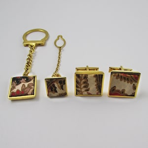 以下が含まれることがあります： キーホルダー、ネクタイピン、カフスボタンを含む、金色のアクセサリーのセット。 各ピースは、ベージュ、茶色、赤の色合いの模様の生地インサートを備えた正方形のデザインが特徴です。 アクセサリーは白い表面に表示されています。