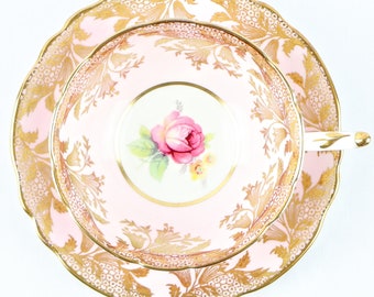 May include: A pink teacup and saucer with a gold floral pattern and a single pink rose in the center. The saucer has a scalloped edge.