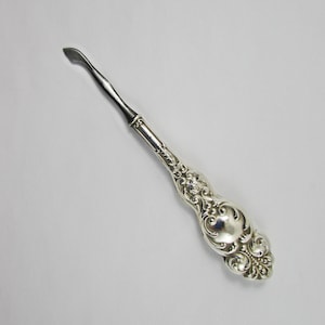May include: A silver nail file with an ornate, detailed handle. The file has a pointed tip and a curved handle.