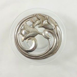May include: A clear glass dish with a silver floral design. The silver design features stylized flowers and leaves in a circular arrangement. The dish is round and sits on a white surface.