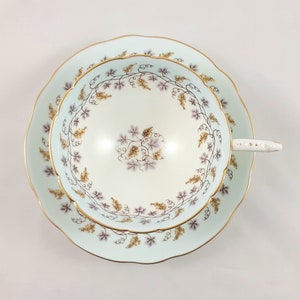 May include: A delicate teacup and saucer set with a pale blue background and a floral pattern of vines and leaves in shades of purple and gold. The set has a gold trim around the edges.