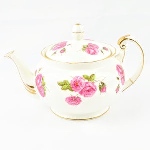 Peut inclure: Une théière blanche avec une bordure dorée et un motif floral de roses roses. La théière a un couvercle avec un bouton sur le dessus.