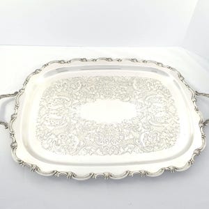 May include: A rectangular silver-toned serving tray featuring ornate, embossed floral and scrollwork patterns across the surface. The tray includes two decorative handles on the sides and a scalloped, raised edge with intricate detailing throughout.