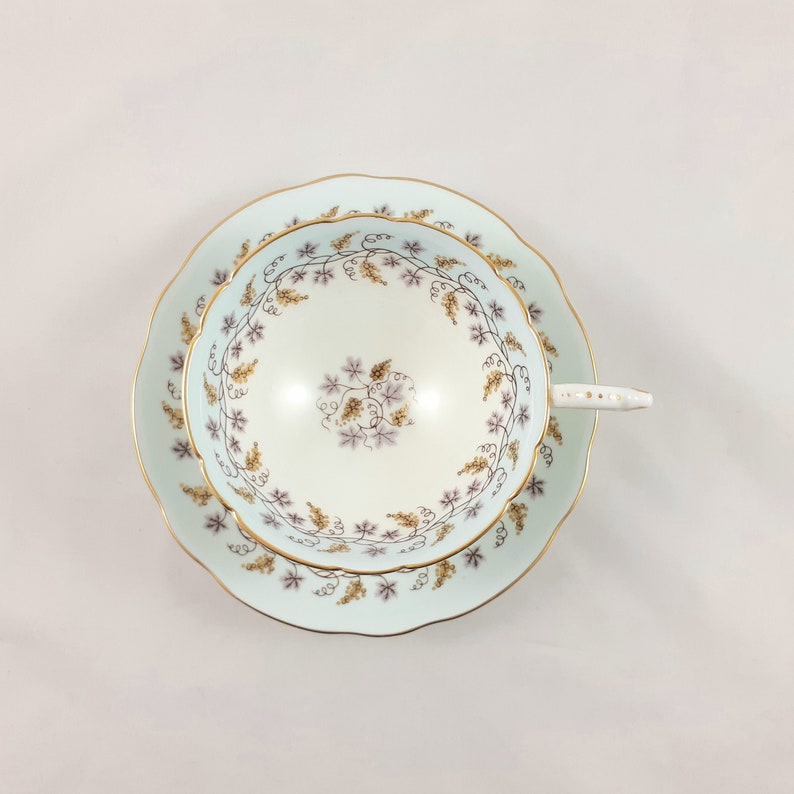 May include: A delicate teacup and saucer set with a pale blue background and a floral pattern of vines and leaves in shades of purple and gold. The set has a gold trim around the edges.