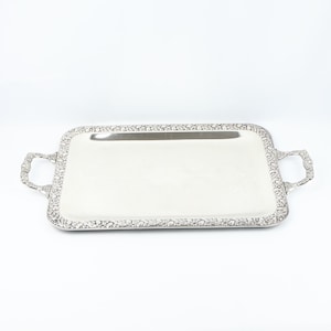 May include: A rectangular silver serving tray with decorative floral edging and handles. The tray has a polished, reflective surface, suitable for serving food or displaying items. The handles are curved and feature the same floral pattern.