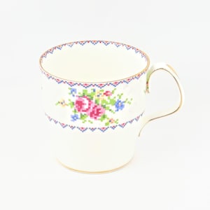 Peut inclure: Tasse blanche avec un motif floral au point de croix. Le motif comprend des fleurs roses, bleues et jaunes. Le bord et la poignée sont dorés. La tasse a une bordure géométrique bleue et rouge.