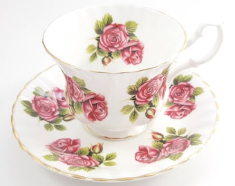 May include: A white teacup and saucer set with a pink rose floral pattern. The set has a gold trim around the edges.