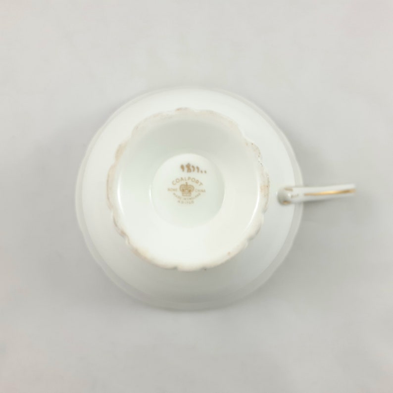 May include: White porcelain teacup with a gold rim and a gold Coalport logo on the bottom. The logo includes a crown and the text "Coalport Fine China England Est. 1750".