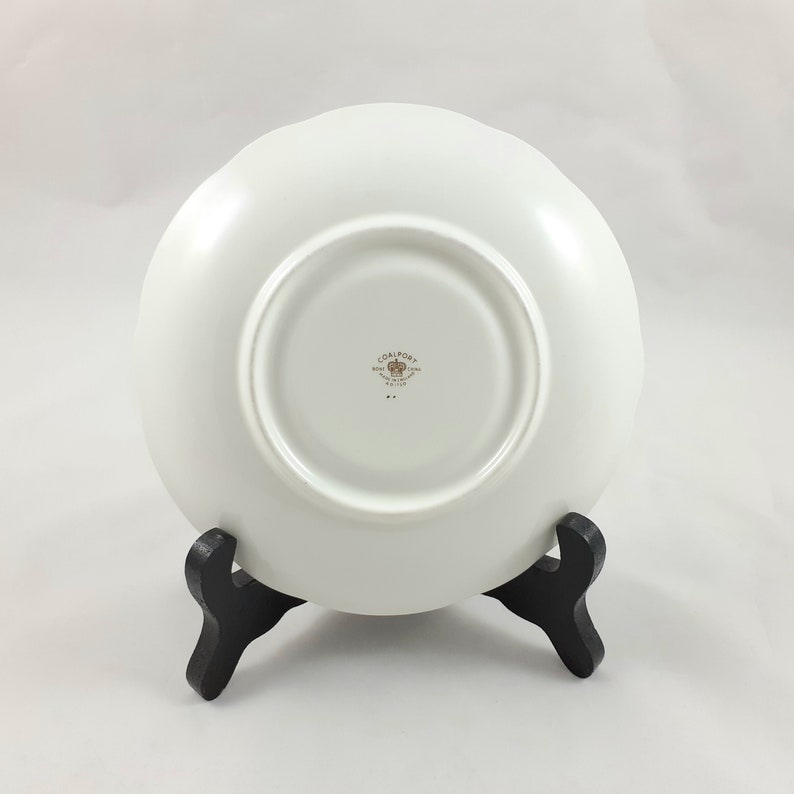 May include: White ceramic saucer with a gold Coalport logo and a gold crown. The saucer is on a black stand.