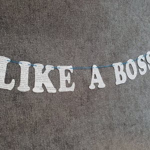 May include: A white paper banner with the text "LIKE A BOSS" strung on a blue cord against a gray textured background.