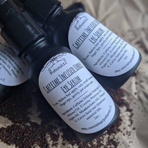 May include: Two black bottles of caffeine infused eye serum with white labels. The labels have the text "PotionsLLC Caffeine Infused Under Eye Serum" and list the ingredients and instructions for use. The bottles are sitting on a white surface with brown coffee grounds.
