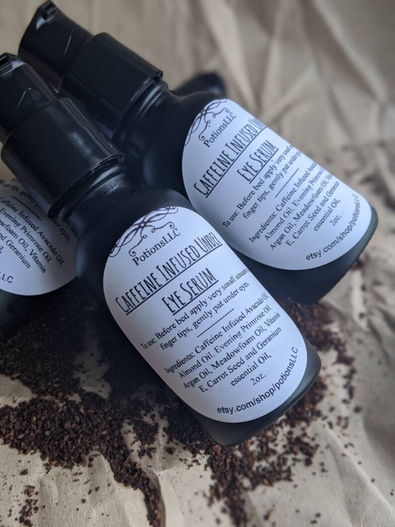 May include: Two black bottles of caffeine infused eye serum with white labels. The labels have the text "PotionsLLC Caffeine Infused Under Eye Serum" and list the ingredients and instructions for use. The bottles are sitting on a white surface with brown coffee grounds.