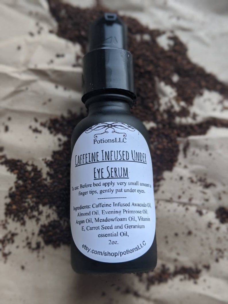 May include: A black glass bottle with a white label that reads "Caffeine Infused Under Eye Serum" and lists the ingredients. The bottle is sitting on a brown surface with coffee grounds scattered around it.