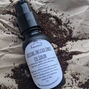 May include: A black bottle with a white label that reads "Caffeine Infused Under Eye Serum" and lists the ingredients. The bottle is sitting on a brown surface with coffee grounds.