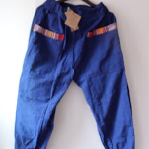 Pode incluir: Calças azuis com detalhes coloridos listrados nos bolsos. As calças estão penduradas em um cabide de madeira. Elas têm cós elástico e um ajuste folgado. Fundo branco.