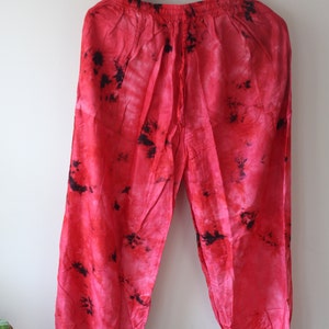 Pode incluir: Calças vermelhas tie-dye com cintura com cordão, penduradas num cabide preto. As calças têm um ajuste folgado e apresentam manchas pretas por todo o tecido vermelho. As calças são feitas de um material leve.
